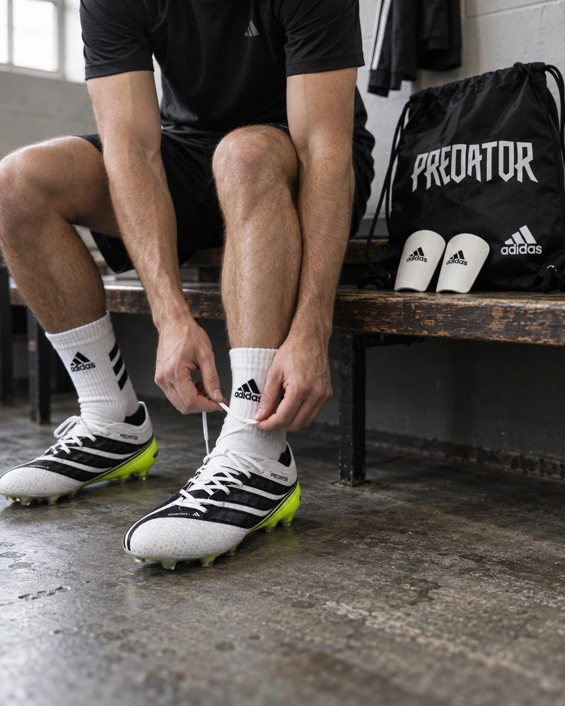 Spieler schnürt seine Adidas Predator Fußballschuhe; Schuhbeutel und Schienbeinschoner liegen auf der Umkleidebank.